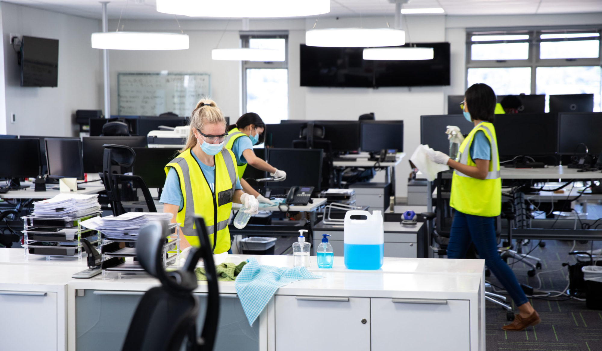 Caucasian woman cleaning an office wearing hi vis vest, gloves and face mask, using disinfectant, colleagues working in background. Hygiene in workplace during Coronavirus Covid 19 pandemic.